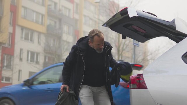 A handsome middle-aged man opens a trunk of his car, puts a backpack and clothes in, and closes the trunk shut