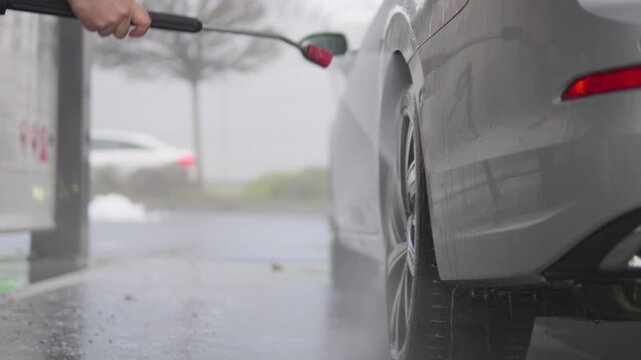 A man power washes the rear wheel and side of his car - low view from behind the car