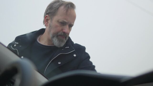 A handsome middle-aged man wipes a windshield with a cloth - view from behind the windshield inside the car