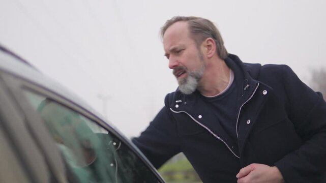 A handsome middle-aged man wipes a windshield with a cloth - view from the side of the car