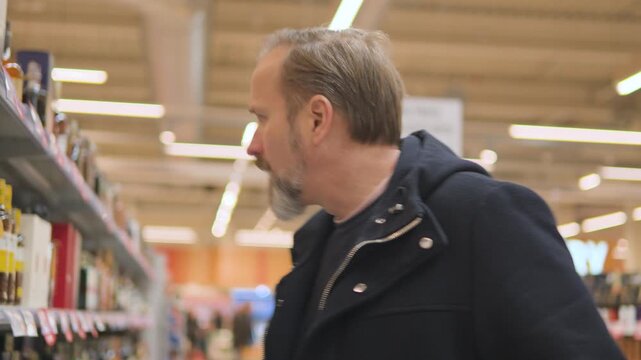 A handsome middle-aged man shops in the liquor section of a supermarket, hesitates, then takes a bottle