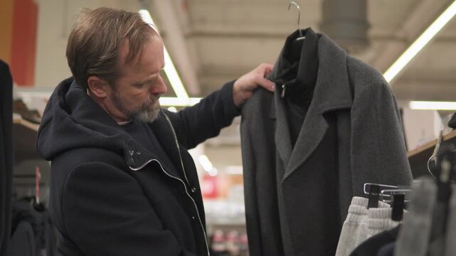 A handsome middle-aged man examines a coat and peels it off the hanger in a supermarket