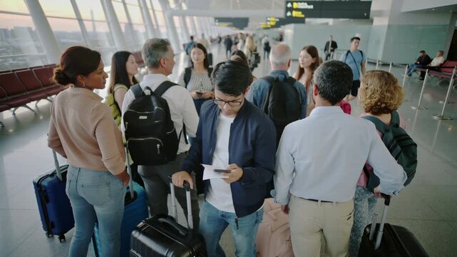 Young man and diverse group of travelers waiting in line at an airport terminal at sunset, checking smartphones and boarding passes for international flight and travel