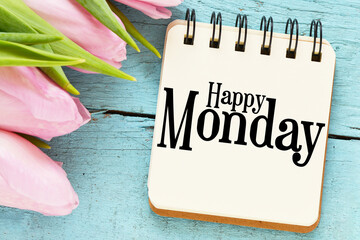 Happy Monday words in a notebook next to tulips on a blue table.