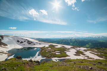 Scenic sunlit landscape with alpine lake in rocky snowy cirque near stone hill top in sunny day during thaw. Ice floats in mountain lake among rocks with view to forest mountain range under bright sun © Daniil