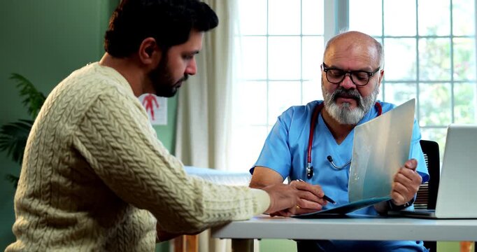 Indian male doctor discussing prescription with young male patient while writing medical advice as experienced physician explains treatment plan inside modern Indian clinic OPD healthcare visit