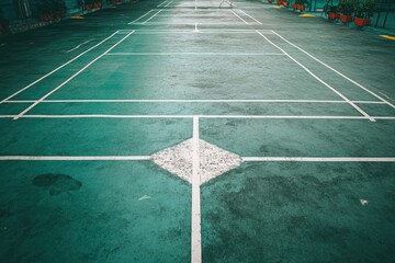 An outdoor sports court, likely for badminton or a similar racket game, showcasing crisp white boundary lines against a weathered, textured teal-green surface. The expansive, empty court evokes a sens