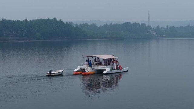 A small tourist catamaran cruises across calm tropical backwaters, towing a motor dinghy and carrying kayaks. Palm trees and a quiet shoreline create a peaceful travel atmosphere.