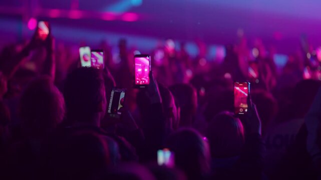 Slow motion crowd jumping dancing and filming live concert as bright stage lights flicker and amplify collective energy