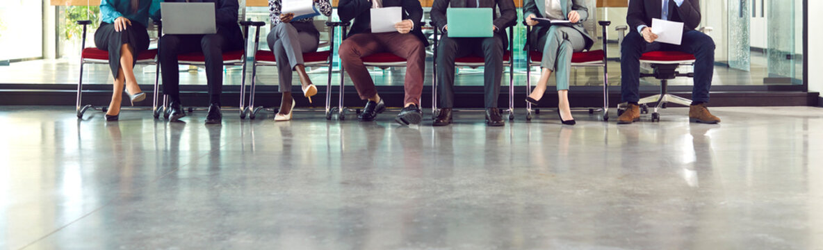 Legs of unrecognizable business people group sitting on chairs in a row with resumes and laptops in their hands. Job candidates seekers waiting for interview invitation turn. Banner.
