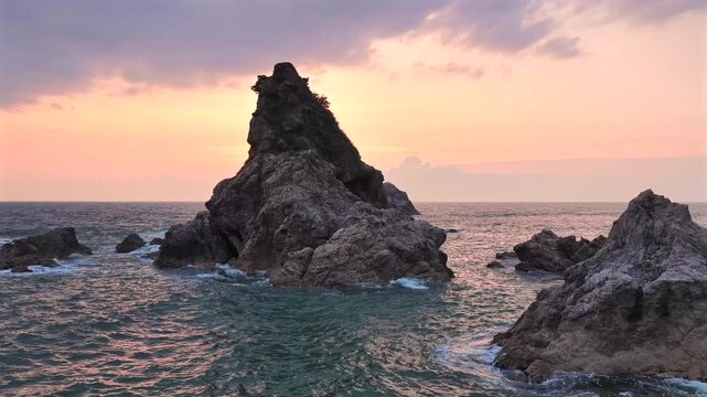 Aerial view of a rugged, dark rocky outcrop piercing the horizon, surrounded by the calm, turquoise sea at sunset, Sendai, Miyagi, Japan.