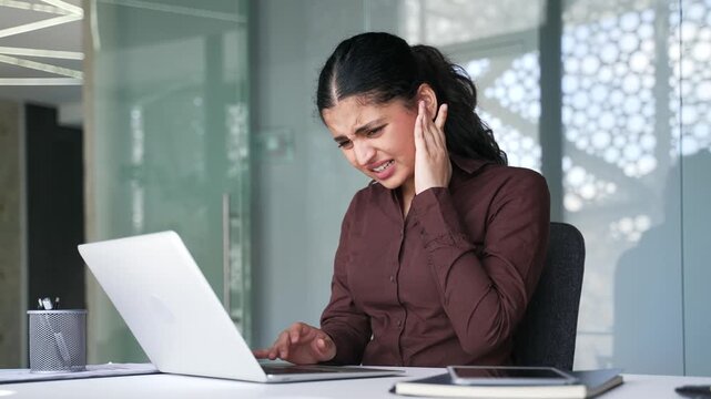 Businesswoman suffers from ear pain while sitting at workplace in office. Upset woman massages her ear with her fingers. She has furuncle, shingles, eczema, ear diseases or otitis media. Earache
