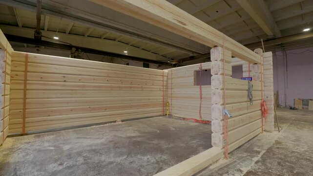 Partially assembled wooden cabin structure inside a workshop, with numbered timber logs, window cutouts, tools, and straps visible. Interior space offers copy space
