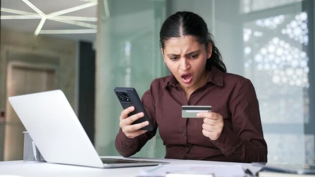 Shocked businesswoman detected fraud checking credit card on phone sitting at workplace in business office. Upset frustrated female became a victim of deception, money was stolen from her account. 