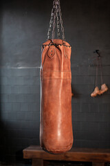Close-up of leather punching bag with metal chains against black wall, rugby boots and wooden bench in gym