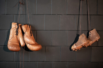 Leather boxing gloves and retro rugby boots hang on a black wall in a gym