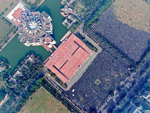 Dhaka, Bangladesh - 31 December 2025: Aerial view of a vast gathering near the National Parliament House, where the red-tiled roofs contrast with the dark waters and surrounding green fields.