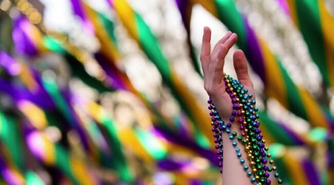 Outstretched hand adorned with vibrant Mardi Gras beads in purple, green, gold, and pink, set against a blurred festive background.