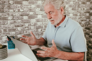 Senior man sitting at desk in home office, looking stressed and frustrated while using laptop. Concept of technology problems, online difficulties, stress, remote work, aging and modern lifestyle