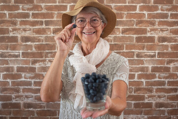 Smiling senior woman holding a glass of fresh blueberries. Healthy eating concept, positive aging, nutrition, and wellbeing, with a brick wall background