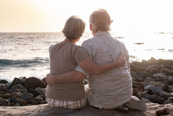 Back view of a bonding senior couple hugging while sitting on a rocky beach face the sea looking at the horizon at sunset light. Retirement lifestyle, vacation, freedom