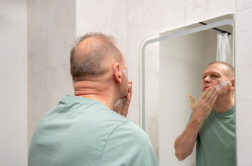A mature man applies shaving foam or skincare product to his face while looking at his reflection in