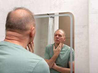 Fototapeta premium A mature man checks his face while looking at his reflection in a bathroom mirror at home. Calm male