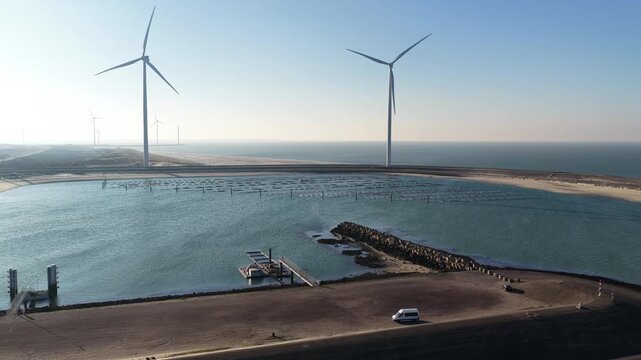 Aerial view of a coastal landscape featuring wind turbines standing tall against the horizon and a small white van parked nearby, Vrouwenpolder, Zeeland, Netherlands.