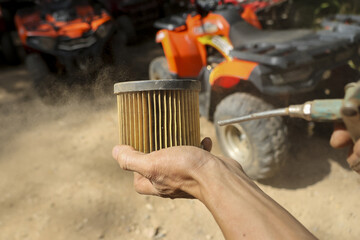 Dirty air filter held in mechanic hand for cleaning maintenance in front of orange ATV vehicle on dusty ground outdoors showing serious concern for engine health and performance checkup