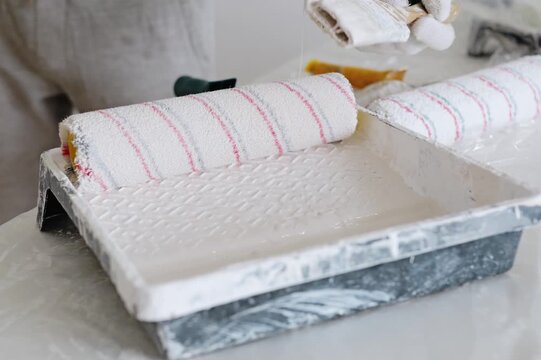 A person pours white paint into a tray and uses a brush while preparing for painting in a home workspace. Rollers sit beside the tray ready for use