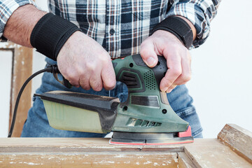 Craftsman. Adult carpenter using an electric sander to smooth an old wooden window. Construction industry, carpentry, housework do it yourself. Restoration.