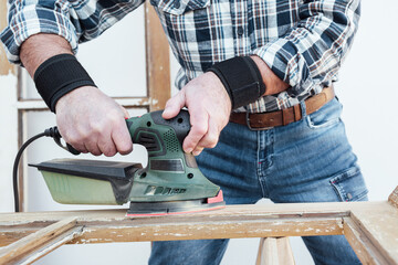Craftsman. Adult carpenter using an electric sander to smooth an old wooden window. Construction industry, carpentry, housework do it yourself. Restoration.