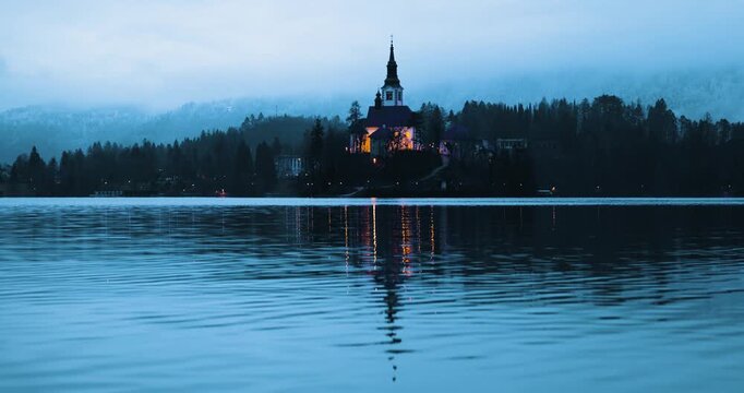 Mari&auml; Himmelfahrt Kirche auf der Insel im Bleder See w&auml;hrend der blauen Stunde, ruhige Wasserfl&auml;che mit dezenter Bewegung und Spiegelung, beleuchtete Kirche bei D&auml;mmerung in Slowenien
