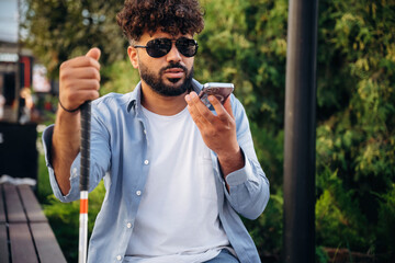 Communication, holding smartphone. Blind man with white cane and sunglasses is in the city