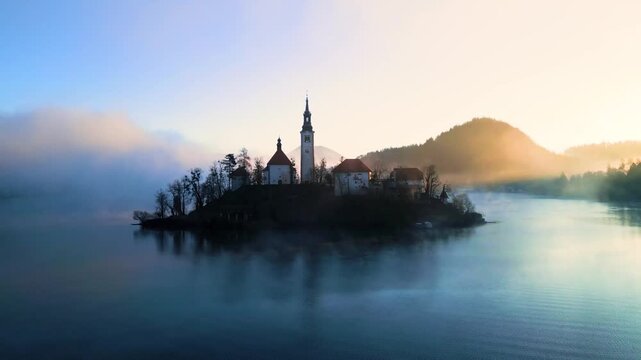 Mari&auml; Himmelfahrt Kirche auf der Insel im See Bled bei Sonnenaufgang mit goldenem Nebel und sanfter Bewegung &uuml;ber Wasser und Landschaft, beleuchtete Kirche im warmen Morgenlicht in Slowenien