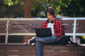 On the bench with laptop on legs. Male student is in the public park outdoors