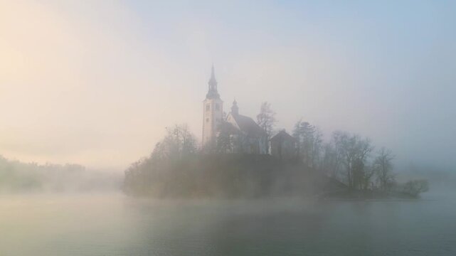 Mari&auml; Himmelfahrt Kirche auf der Insel im See Bled bei Sonnenaufgang, goldener Nebel &uuml;ber dem Wasser, gegen&uuml;berliegende Uferreihe verschwinden im Dunst, Sonnenstrahlen brechen durch den Nebel
