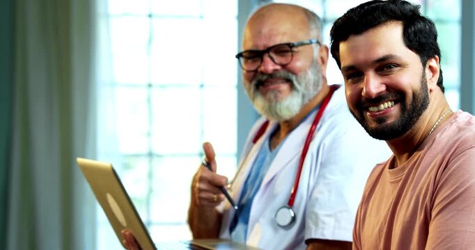 Indian doctor patient doing thumbs up together while using laptop after consultation as experienced male physician and young man celebrate recovery inside modern Indian clinic OPD healthcare visit