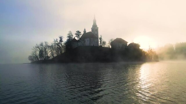 Mari&auml; Himmelfahrt Kirche auf der Insel im See Bled bei Sonnenaufgang, goldener Nebel &uuml;ber dem Wasser, gegen&uuml;berliegende Uferreihe verschwinden im Dunst, Sonnenstrahlen brechen durch den Nebel