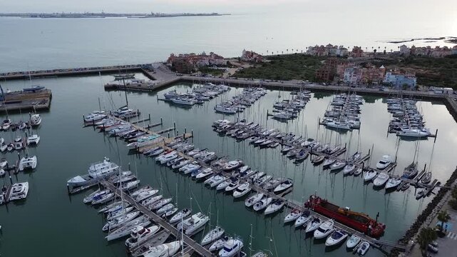 A 4K aerial drone shot ascending and slowly pulling back over Puerto Sherry in C&aacute;diz, Spain. The footage reveals a wide aerial view of the marina with numerous boats docked below, while the city of