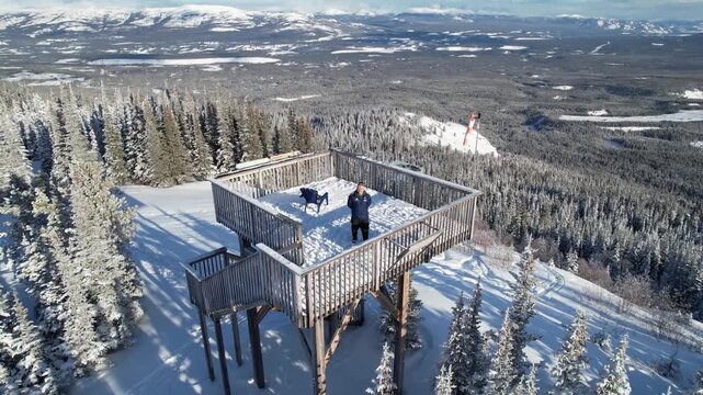 Pulls out from Charles Huang on an observation deck to reveal the vast, snow-covered landscape and nearby mountains near Mount Sima, Yukon
