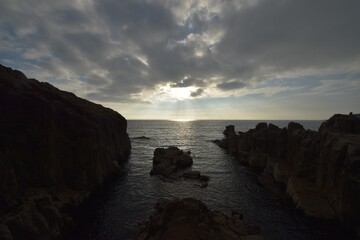A dramatic silhouette of the rugged columnar basalt cliffs of Tojinbo in Fukui Prefecture, Japan, set against a moody sky with sunlight breaking through the clouds over the Sea of Japan.