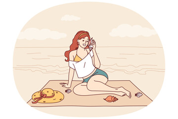 Woman sits on beach and listens to sound of sea from shell, enjoying summer vacation at tropical resort. Tourist girl sunbathes on beach of paradise island in ocean during weekend.