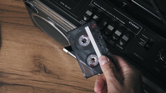 High angle view of a hand flipping and re-inserting a transparent cassette into a retro ghettoblaster. The inner reels and wood grain background highlight the analog aesthetic.