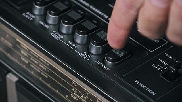 Angled close-up of a finger pressing the record button on a vintage tape deck. The silver trimmed keys and radio tuner scale are visible in the shallow depth of field.