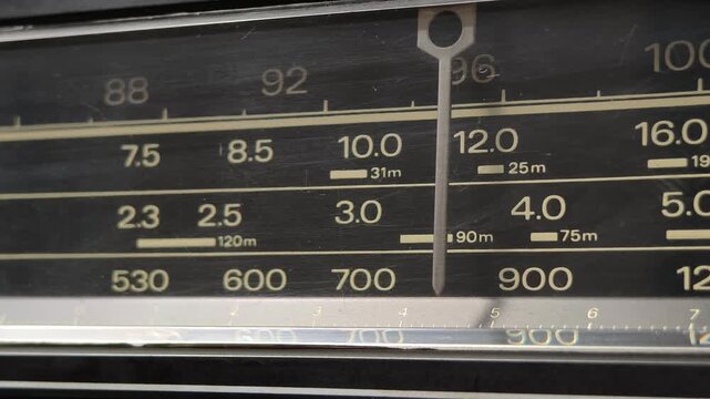 Extreme close-up of an old radio tuner display with the indicator needle scanning a scratched scale. The AM and shortwave bands on the black background emphasize the vintage age.