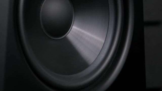 Angled close-up of a black speaker woofer membrane pulsing powerfully in slow motion. The dynamic driver creates intense low-frequency vibrations in a music studio.