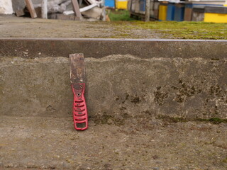 Metal putty knife with worn blade and red handle leaning against a concrete curb.
