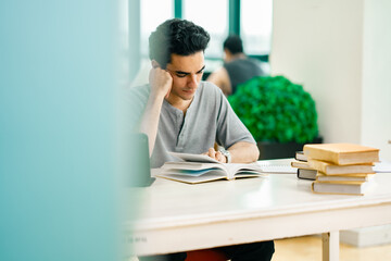 Young man reading an open book while studying at a library desk with a tablet nearby. The image reflects academic focus, modern education, and independent learning.