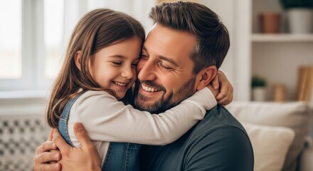 A smiling Caucasian father in a grey t-shirt embraces his cheerful young daughter in denim overalls indoors at home.